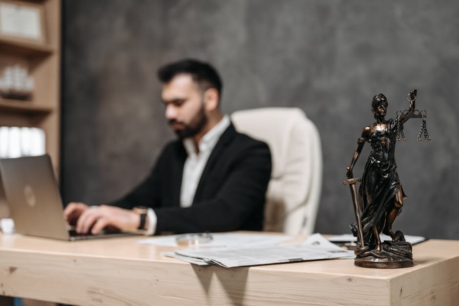 Justice Shekhar Yadav Retires from Allahabad High Court Amid Impeachment Proceedings A businessman working on a laptop in an office with a Lady Justice statue in focus on the desk.