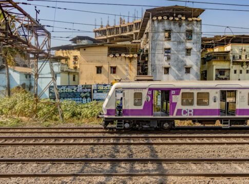 A colorful electric train travels through the urban landscape of Mumbai, India, showcasing vibrant architecture.