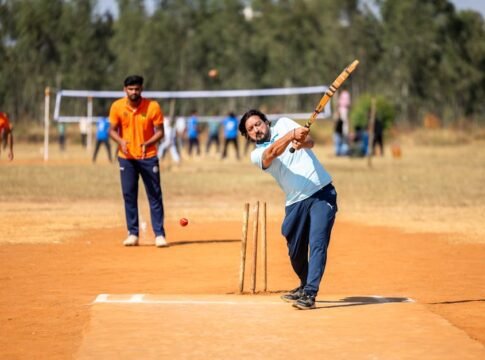 A cricket player in mid-swing during an outdoor match on a sunny day.