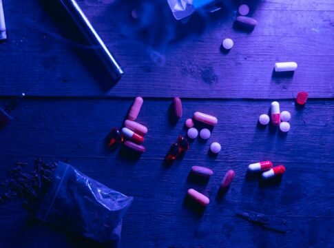 A flat lay of various drugs including pills, capsules, and a syringe on a wooden surface under moody blue lighting. A flat lay of various drugs including pills, capsules, and a syringe on a wooden surface under moody blue lighting.
