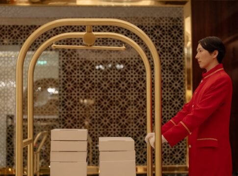 A hotel bellboy in uniform pushes a luggage cart with boxes indoors, demonstrating hospitality service. A hotel bellboy in uniform pushes a luggage cart with boxes indoors, demonstrating hospitality service.