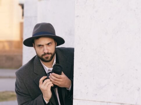 A man in a hat peeks around a corner holding a camera, suggesting secrecy or espionage.