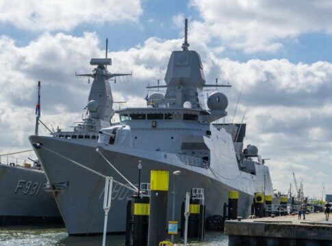A modern naval warship moored at the port of Rotterdam under a blue sky with clouds.