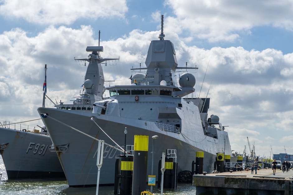 A modern naval warship moored at the port of Rotterdam under a blue sky with clouds.