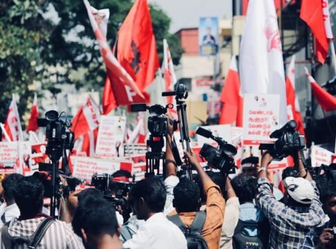 A vibrant protest in Chennai, India with media coverage featuring bright red flags and banners.