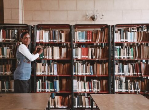 A woman stands beside tall bookshelves filled with books in a library setting.