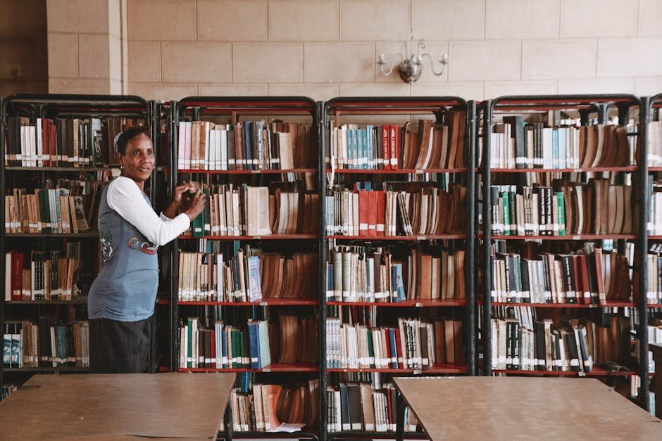 A woman stands beside tall bookshelves filled with books in a library setting.