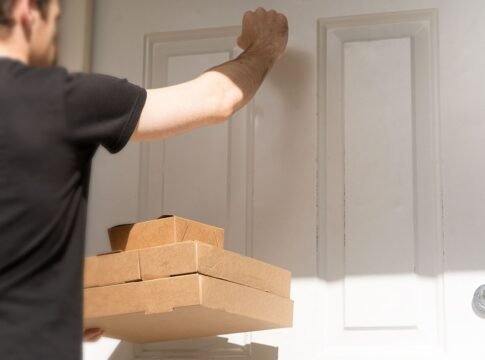 An image of a delivery driver knocking on a white front door to deliver three cardboard takeaway boxes. This image is free to use under the Creative Commons Attribution Licence. Feel free to use it w…