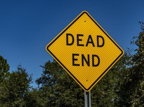 Close-up of a yellow 'Dead End' sign against a blue sky and trees, symbolizing caution and warning.