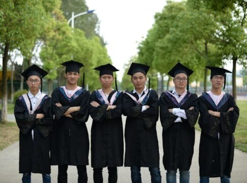 Group of young graduates in gowns and caps standing outdoors, celebrating academic achievement.