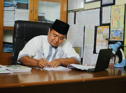 Indonesian educator writing at his desk, focused on paperwork in an office setting.