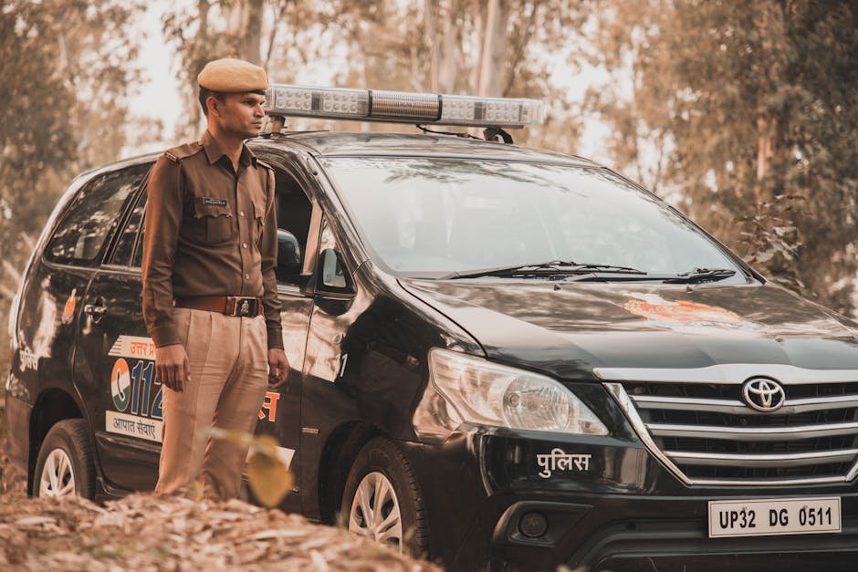 Police officer in uniform standing beside a parked Toyota Innova police vehicle in a wooded outdoor area.