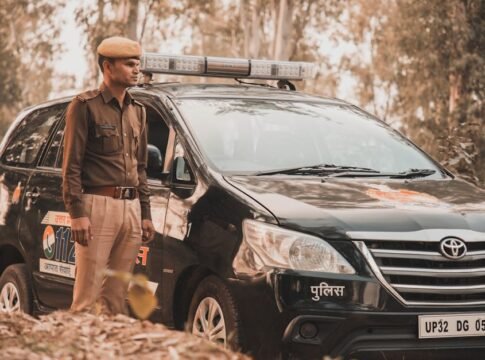 Police officer in uniform standing beside a parked Toyota Innova police vehicle in a wooded outdoor area.