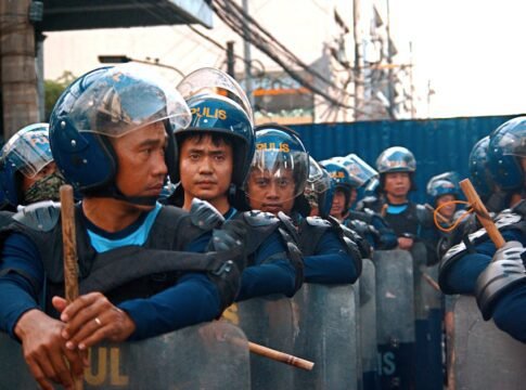 Riot police officers lined up with helmets and shields, maintaining order in a city setting.
