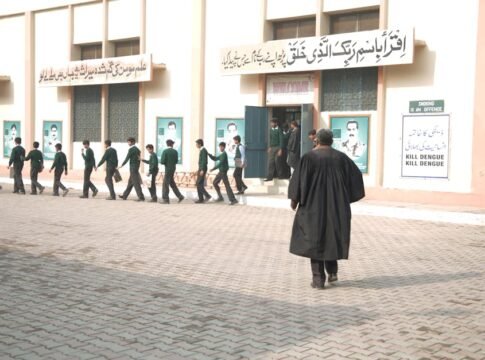Students in uniform lined up at a school entrance with a teacher overseeing.