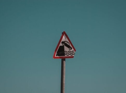 Triangular traffic warning sign showing a car falling into water, set against a clear blue sky in Dublin, Ireland.