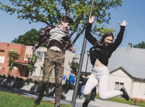 Two teenagers joyfully jumping outdoors in Šiluva, Lithuania under a bright blue sky. Captured in springtime.