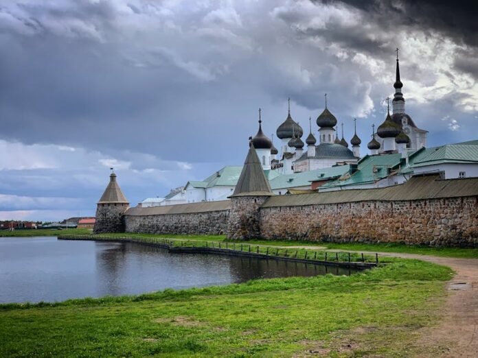 View of Solovetsky Monastery with its iconic domes reflecting on a calm river under a cloudy sky. View of Solovetsky Monastery with its iconic domes reflecting on a calm river under a cloudy sky.