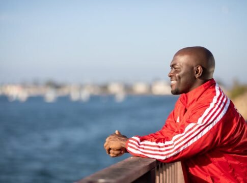 A bald man in a red jacket smiles while leaning on a railing overlooking a blurred ocean background.