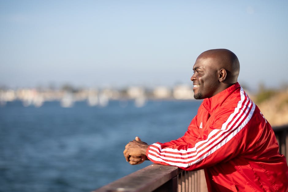 Jason Collins, NBA’s First Openly Gay Player, Dies at 47 A bald man in a red jacket smiles while leaning on a railing overlooking a blurred ocean background.