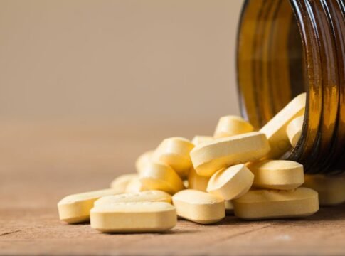 A close-up image of vitamin tablets spilling out of a transparent brown bottle on a wooden surface. A close-up image of vitamin tablets spilling out of a transparent brown bottle on a wooden surface.