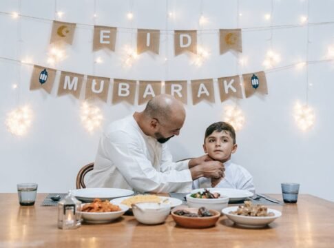 A father adjusts his son's shirt as they sit together celebrating Eid indoors with festive lights and food.