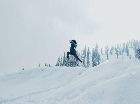 A man leaps across a snow-covered hillside against a backdrop of trees in Gulmarg.