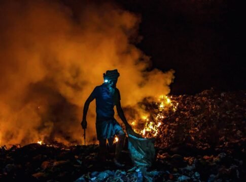 A silhouette of a person working amidst burning garbage piles at night, creating a dramatic scene.