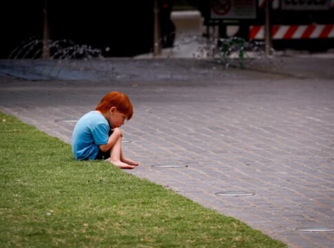 A young red-haired child sits thoughtfully on the pavement near a grassy area in an urban setting. A young red-haired child sits thoughtfully on the pavement near a grassy area in an urban setting.