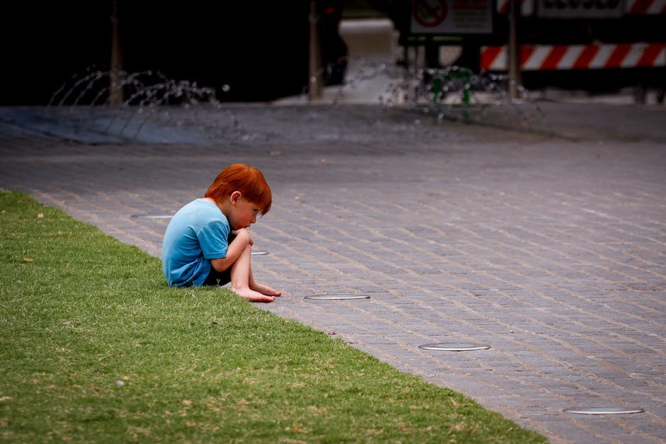 Jammu and Kashmir: 12-Year-Old Girl Found Dead in Budgam Field A young red-haired child sits thoughtfully on the pavement near a grassy area in an urban setting.