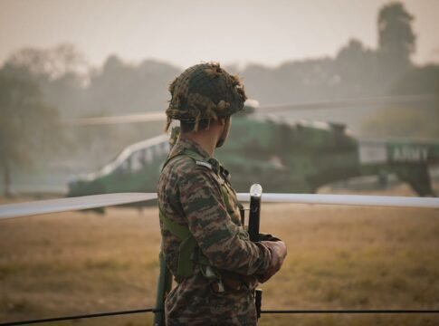An Indian soldier stands guard in a field with a military helicopter in the background.