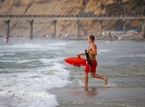 Lifeguard running into the ocean holding a rescue tube at a beach with a pier in the background. Lifeguard running into the ocean holding a rescue tube at a beach with a pier in the background.