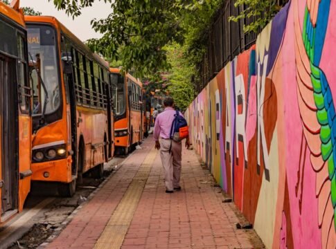 Man walking beside colorful street mural and lined buses in New Delhi, India.