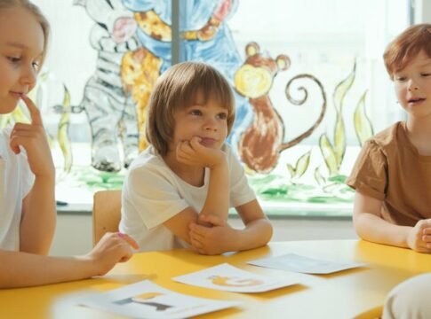 Three children sitting at a yellow table in a classroom, engaging in a learning activity.