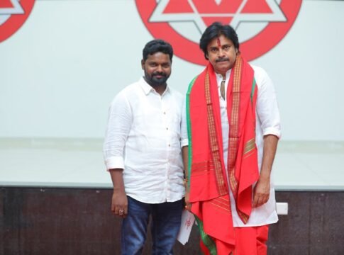 Two Indian men indoors, one in traditional attire, with a logo background.