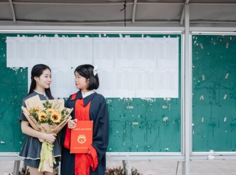 Two graduates celebrate in front of a bulletin board with a sunflower bouquet and diploma.