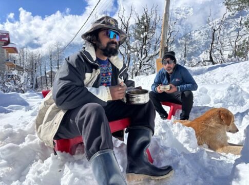 Two men seated in snowy landscape of Manali with a dog, enjoying a meal outdoors amidst a winter wonderland.