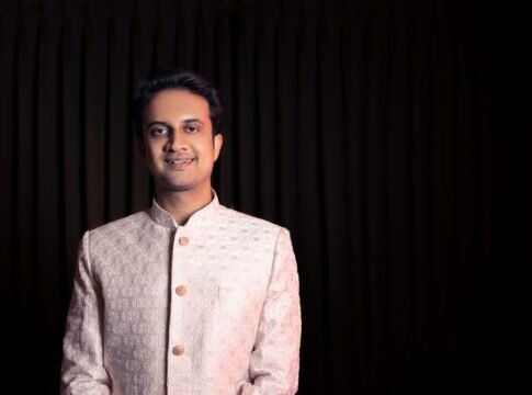 A man in traditional Indian attire posing with a smile against a dark background.