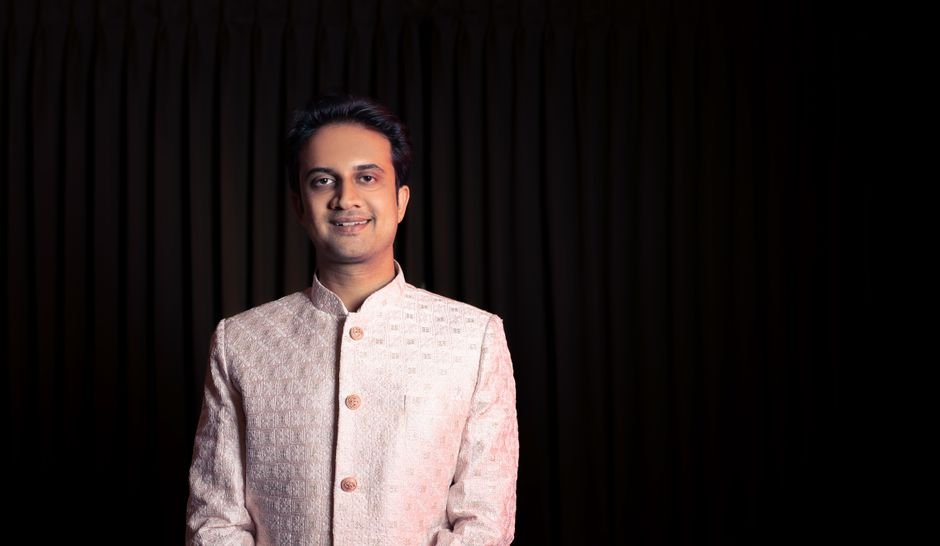 A man in traditional Indian attire posing with a smile against a dark background.
