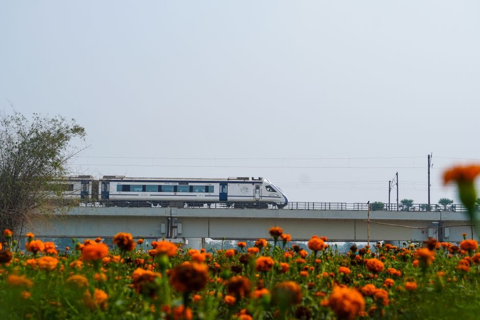 Amritsar-Katra Vande Bharat Express Rerouted Via Batala, Gurdaspur for Faster Journey A sleek high-speed train travels across a raised bridge above vibrant orange marigold flowers.