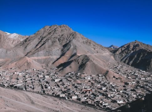 A stunning aerial view of Leh with its rugged mountain backdrop and scattered village buildings under a clear blue sky.