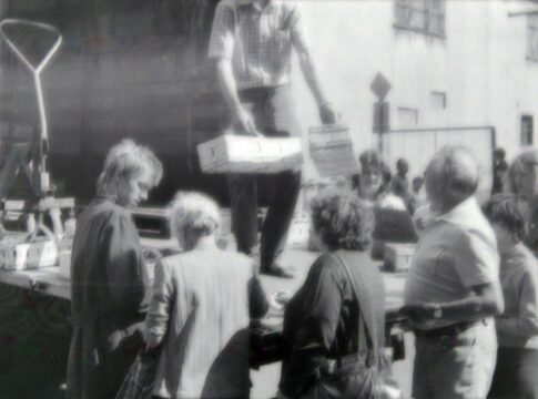 A vintage black and white photo showing people gathering at an outdoor market with goods being unloaded.