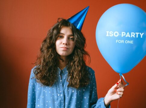 A young woman wearing a party hat holding a balloon that says 'ISO-PARTY FOR ONE.' A young woman wearing a party hat holding a balloon that says 'ISO-PARTY FOR ONE.'