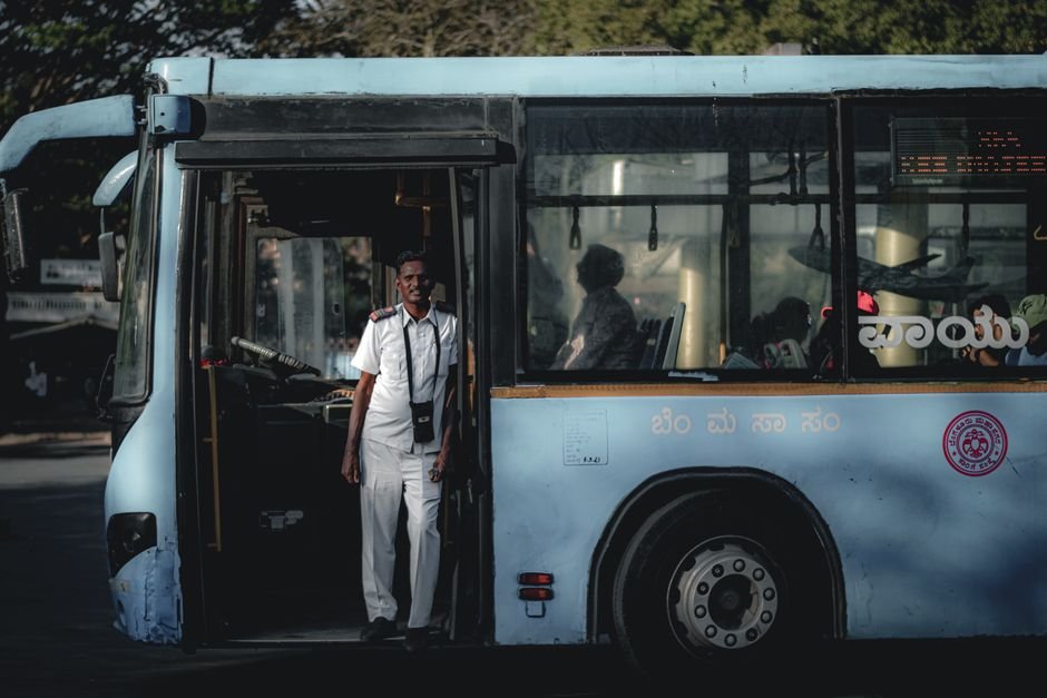 Sivakumar K.B. Assumes Charge as Managing Director of KSRTC in Karnataka Bus driver wearing a white uniform standing by the bus door, sunlight casting shadows inside