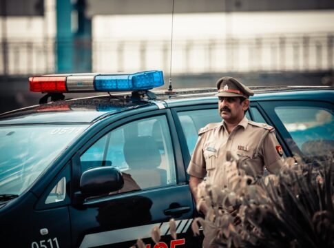 Male in uniform standing near patrol car with flashing lights and looking away