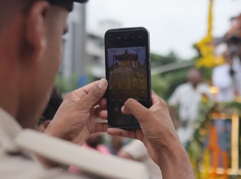 A person photographs the colorful Jagannath Rath Yatra parade with a smartphone.