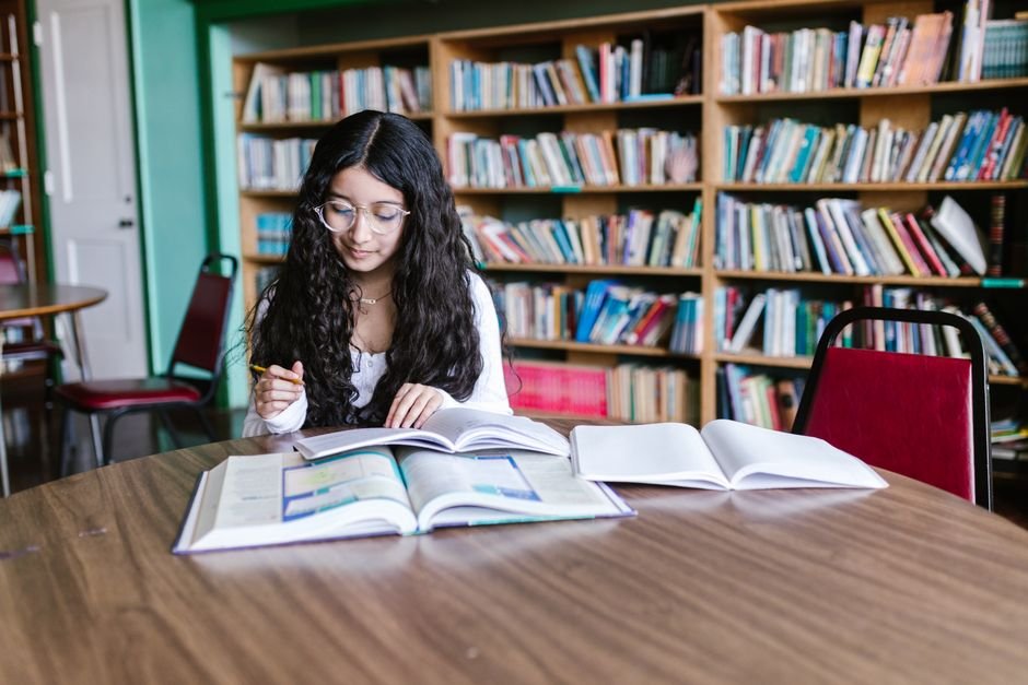Jammu Education Directorate Orders Comprehensive Review of All School Library Books A young woman studying at a library table surrounded by bookshelves, focused on her work.