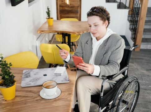 High angle of focused female in wheelchair browsing cellphone at table with netbook and cup of latte High angle of focused female in wheelchair browsing cellphone at table with netbook and cup of latte