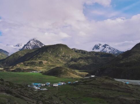 Picturesque village nestled in lush mountains with snow-capped peaks under a cloudy sky.