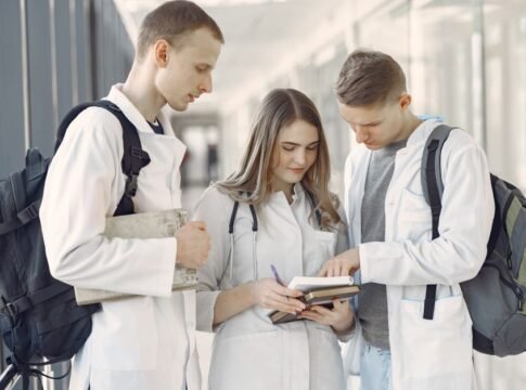 Three medical students in a hallway discussing notes, fostering teamwork and learning. Three medical students in a hallway discussing notes, fostering teamwork and learning.
